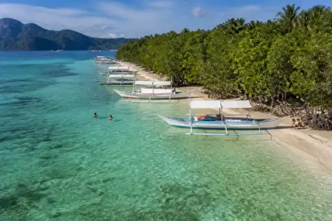 Shallow turquoise water, pale sand and anchored outrigger boats at CYC Beach, Coron