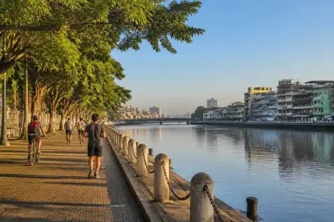 People walking and cycling along a green riverside esplanade in Manila at sunrise