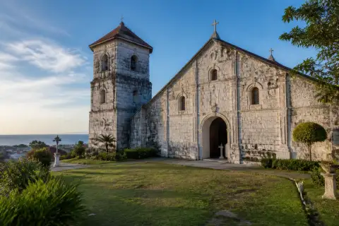 Front façade and bell tower of Boljoon Church with open church grounds and coastal town backdrop
