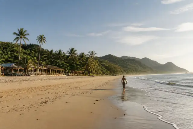 Long open sandy shoreline at Duli Beach with sparse beach structures and a surfer near the water