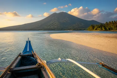 Wooden banca approaching a white sandbar with a volcanic island in the background at golden hour