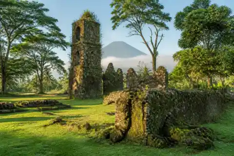 Moss-covered coral-stone walls of the Guiob Old Church ruins with trees and volcanic backdrop