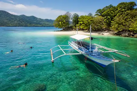 Traditional outrigger boat near a small sand islet off Port Barton with snorkelers in clear water
