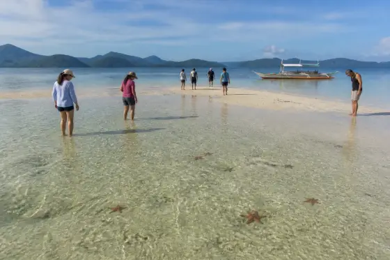 Visitors standing on a shallow sandbar with a small banca anchored offshore at Port Barton