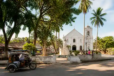 Parish church and shaded town plaza in a Siquijor village with coconut trees and a passing tricycle